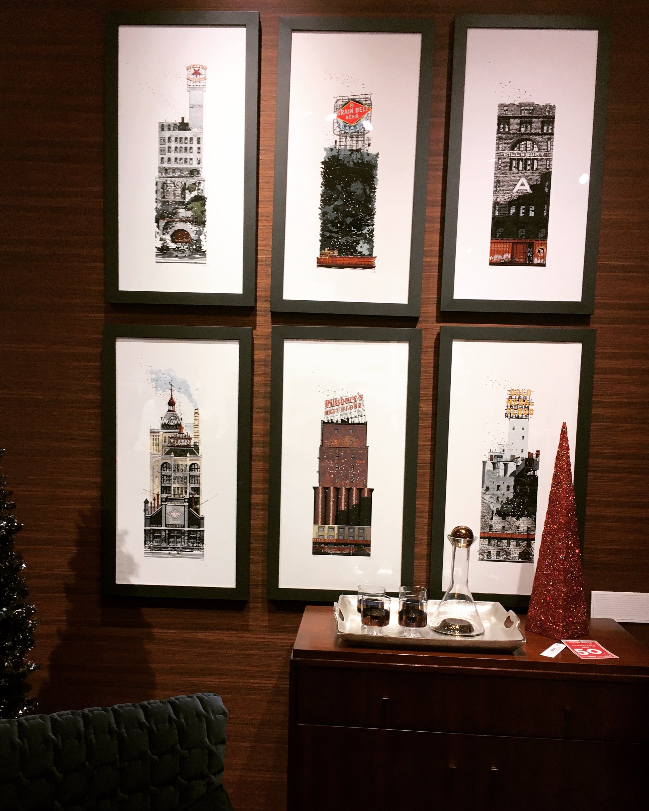Six framed art prints featuring vintage skyscrapers arranged in a 2x3 grid on a dark wood-paneled wall above a wooden cabinet with a tray and a red decorative tree.